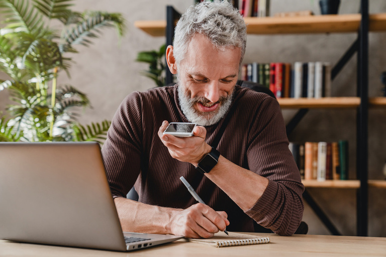 Senior man writing notes in notepad while talking by speakerphone from workspace